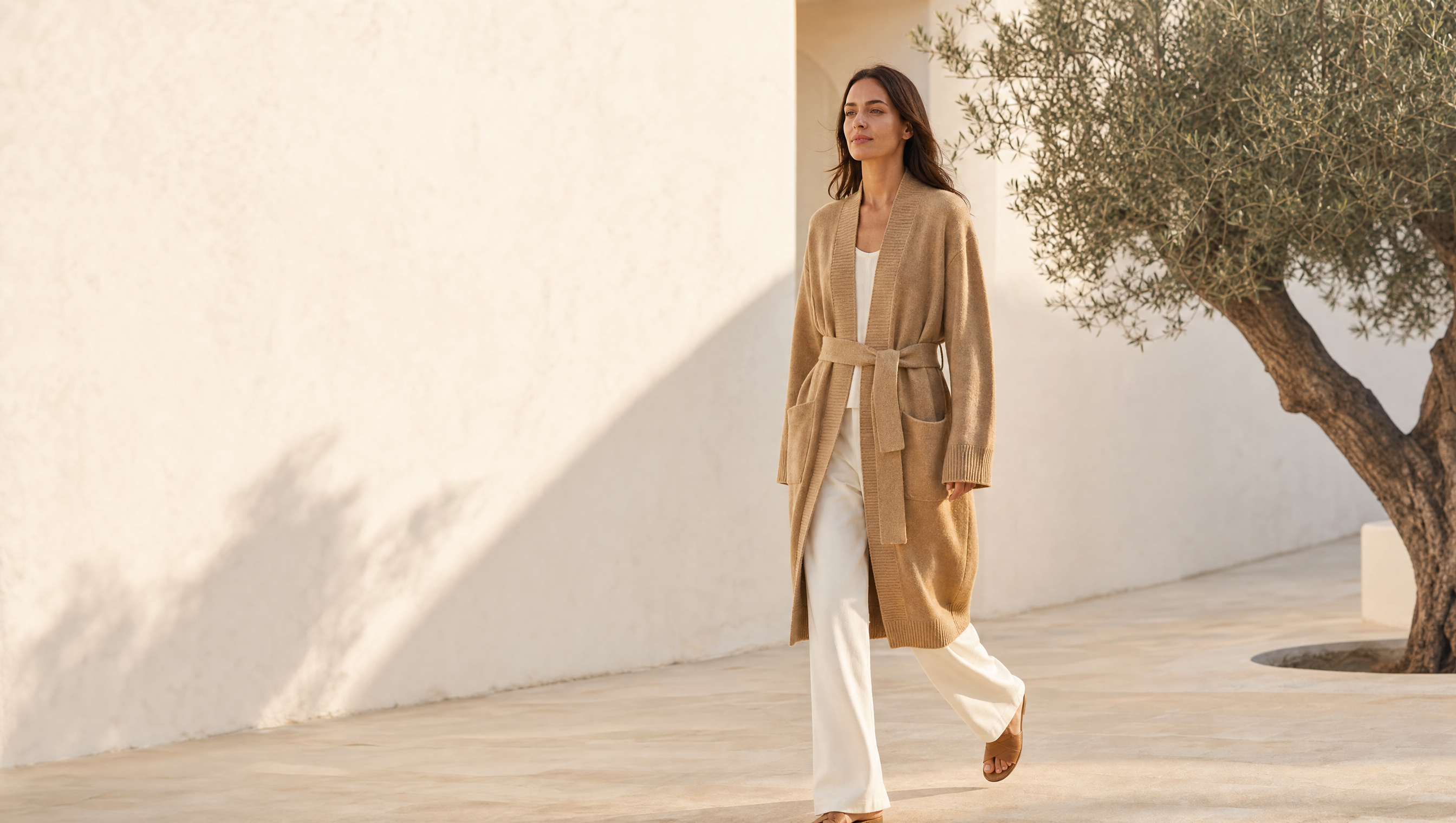 A woman walking outdoors in a beige long cardigan over a white outfit, set against a sunlit white stucco wall and a tree.